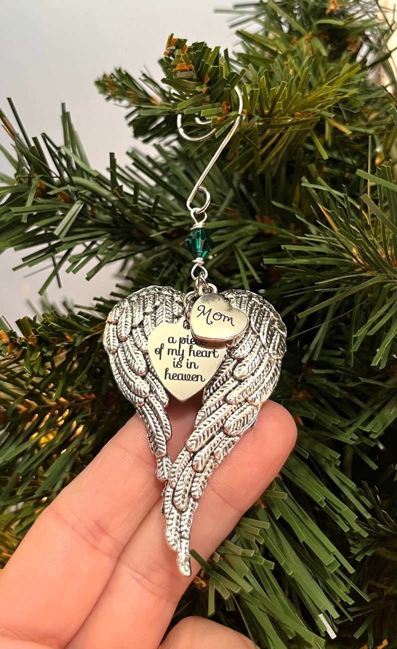 May include: Silver angel wing ornament with a heart that says "a piece of my heart is in heaven" and a smaller heart that says "Mom". The ornament has a green crystal bead.