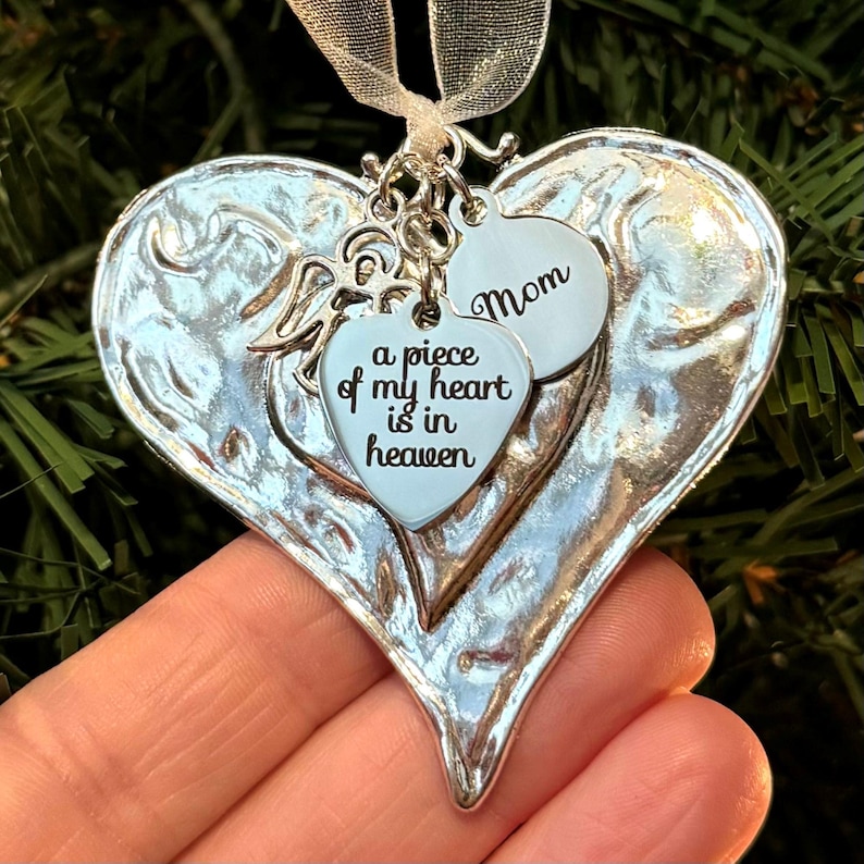 Puede incluir: Adorno plateado en forma de coraz&oacute;n con un acabado texturizado. Presenta un colgante m&aacute;s peque&ntilde;o en forma de coraz&oacute;n con la inscripci&oacute;n "a piece of my heart is in heaven" y un colgante redondo que dice "Mom". Se adjunta una cinta blanca para colgar.