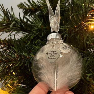 May include: A clear glass Christmas ornament with a white feather inside. The ornament has a silver metal top and a silver chain with two silver heart charms. One charm says "Mom" and the other says "a piece of my heart is in heaven".