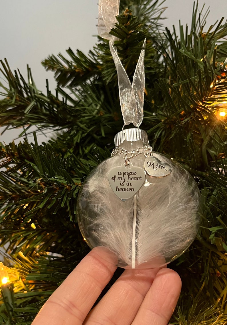 May include: A clear glass Christmas ornament with a white feather inside. The ornament has a silver metal top and a silver chain with two silver heart charms. One charm says "Mom" and the other says "a piece of my heart is in heaven".