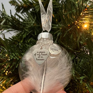 May include: A clear glass Christmas ornament with a white feather inside. It has two silver heart charms attached, one with the text "a piece of my heart is in heaven" and the other with the text "Mom".