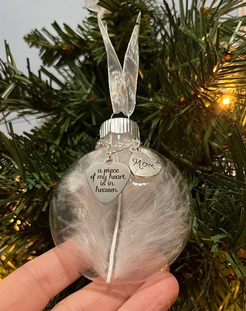 May include: A clear glass Christmas ornament with a white feather inside. It has two silver heart charms attached, one with the text "a piece of my heart is in heaven" and the other with the text "Mom".
