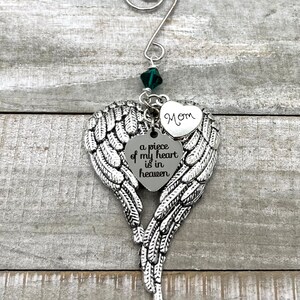 May include: Silver angel wing ornament with a heart-shaped charm that reads "a piece of my heart is in heaven" and a smaller heart charm that reads "Mom". A green crystal hangs from the top of the ornament.