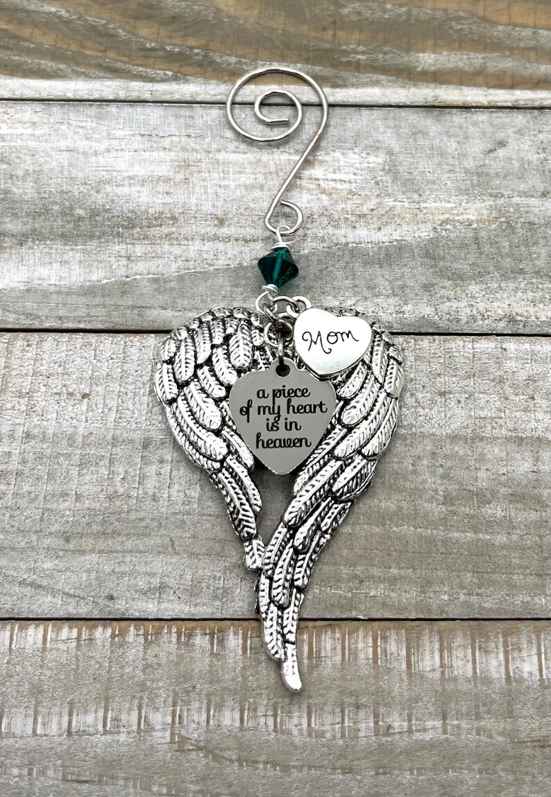 May include: Silver angel wing ornament with a heart-shaped charm that reads "a piece of my heart is in heaven" and a smaller heart charm that reads "Mom". A green crystal hangs from the top of the ornament.