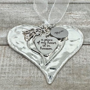 Puede incluir: Adorno plateado en forma de coraz&oacute;n con un coraz&oacute;n m&aacute;s peque&ntilde;o y un dije redondo. El coraz&oacute;n m&aacute;s peque&ntilde;o dice "a piece of my heart is in heaven". El dije redondo dice "Mam&aacute;". Un ala de &aacute;ngel y una cinta blanca est&aacute;n adjuntas.