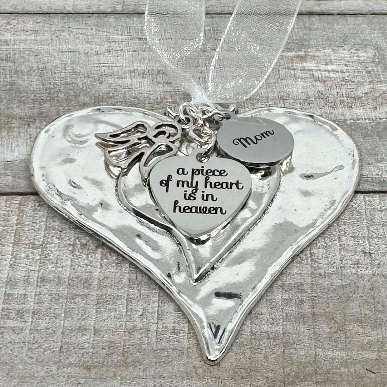 Puede incluir: Adorno plateado en forma de coraz&oacute;n con un coraz&oacute;n m&aacute;s peque&ntilde;o y un dije redondo. El coraz&oacute;n m&aacute;s peque&ntilde;o dice "a piece of my heart is in heaven". El dije redondo dice "Mam&aacute;". Un ala de &aacute;ngel y una cinta blanca est&aacute;n adjuntas.
