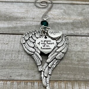 May include: A silver angel wing ornament with a heart charm that reads "a piece of my heart is in heaven" and a smaller heart charm that reads "Mom". The ornament is decorated with a green crystal bead.