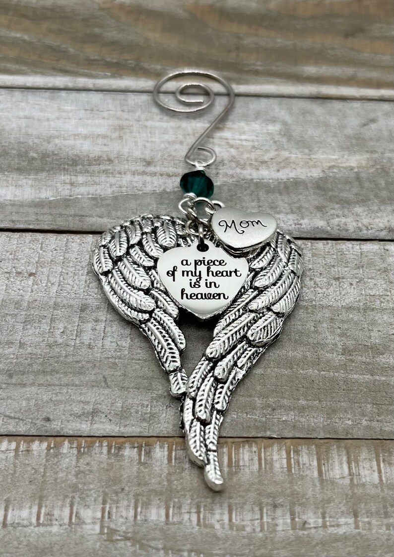 May include: A silver angel wing ornament with a heart charm that reads "a piece of my heart is in heaven" and a smaller heart charm that reads "Mom". The ornament is decorated with a green crystal bead.