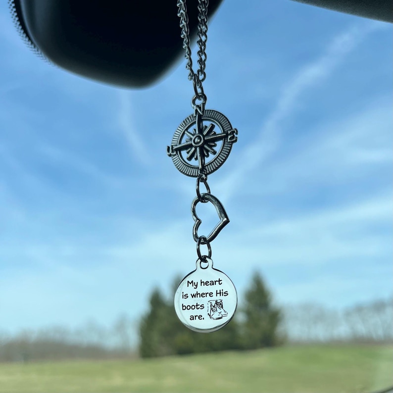 K&ouml;nnte beinhalten: Ein silberner Auto-Anh&auml;nger mit einem Kompass, einem Herzen und einem runden Anh&auml;nger mit der Aufschrift "Mein Herz ist, wo seine Stiefel sind."