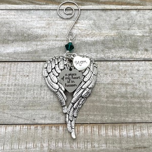 May include: Silver angel wing ornament with a heart charm that reads "a piece of my heart is in heaven" and a smaller heart charm that reads "Mom". The ornament is decorated with a green crystal.