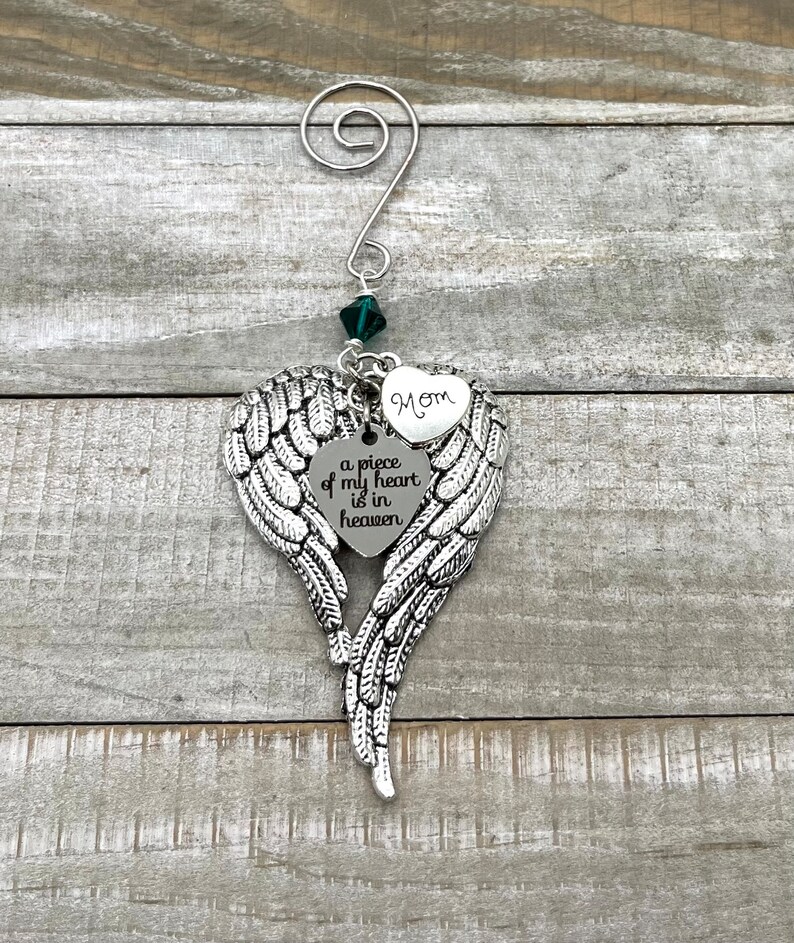 May include: Silver angel wing ornament with a heart charm that reads "a piece of my heart is in heaven" and a smaller heart charm that reads "Mom". The ornament is decorated with a green crystal.