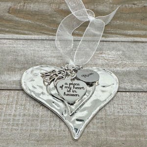 Puede incluir: Adorno plateado en forma de coraz&oacute;n con superficie texturizada, con peque&ntilde;os dijes de coraz&oacute;n y una cinta. Un dije dice "Mom", y otro dice "a piece of my heart is in heaven".