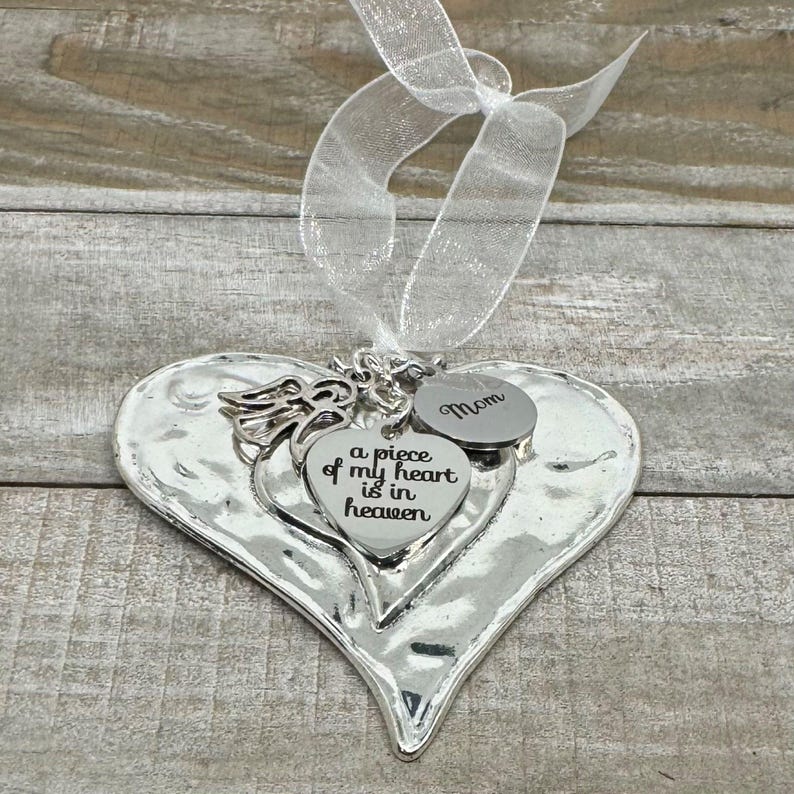 Puede incluir: Adorno plateado en forma de coraz&oacute;n con superficie texturizada, con peque&ntilde;os dijes de coraz&oacute;n y una cinta. Un dije dice "Mom", y otro dice "a piece of my heart is in heaven".