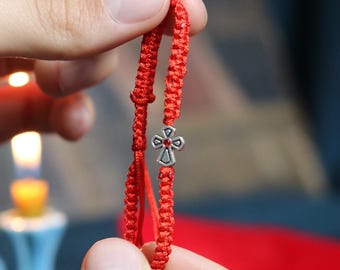 May include: A red woven bracelet with a silver cross charm. The bracelet is held between two hands, with a blurred background featuring a candle and red fabric. The cross has a detailed design.