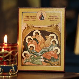 May include: An icon depicting religious figures in various robes, with halos, resting in a cave-like setting. The icon is framed in gold and rests on a dark surface. A lit red candle in a decorative holder is in the foreground.