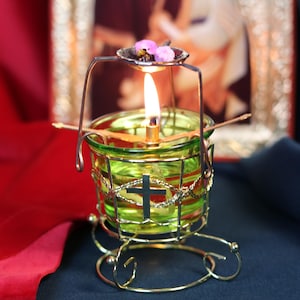 May include: A lit green glass candle holder with a gold metal frame and a cross design. The candle holder has a small dish with pink flowers and is set against a blurred religious icon background.