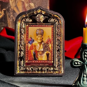 May include: A small, ornate, gold-framed icon depicting a religious figure with a crown and a book. The icon is on a black surface with a lit candle in a green cross-shaped holder.