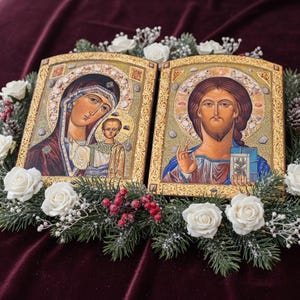 May include: Two religious icons with gold frames, depicting Mary and Jesus, surrounded by a wreath of white roses, greenery, and red berries. The icons are set against a deep burgundy background.