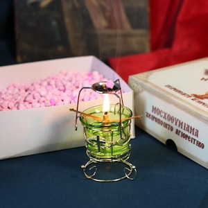 May include: A lit green glass candle holder with a gold metal frame. A small spoon holds incense above the flame. A box of pink granules and a box with Greek text are in the background. The candle holder has a cross design.