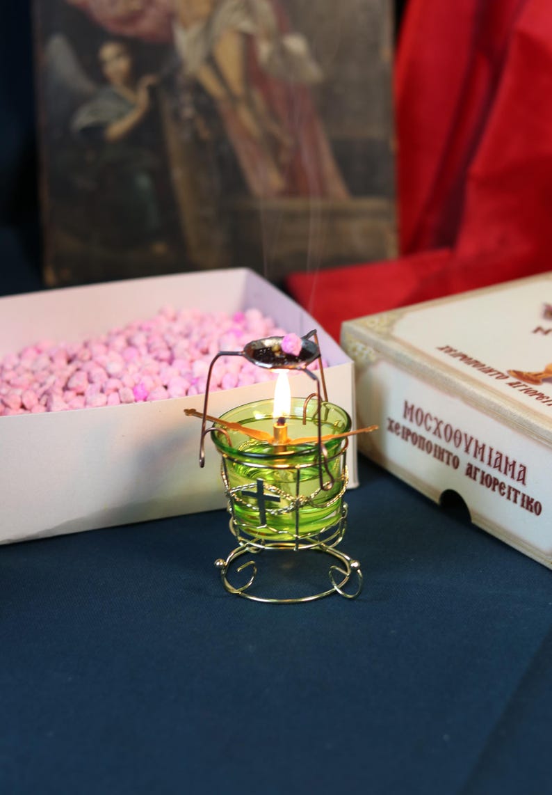 May include: A lit green glass candle holder with a gold metal frame. A small spoon holds incense above the flame. A box of pink granules and a box with Greek text are in the background. The candle holder has a cross design.