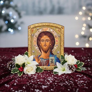 May include: An icon of Jesus Christ with a gold frame, adorned with white roses, lilies, pinecones, and holly berries. The icon is set against a burgundy velvet surface with a snowy effect, creating a festive and religious theme.