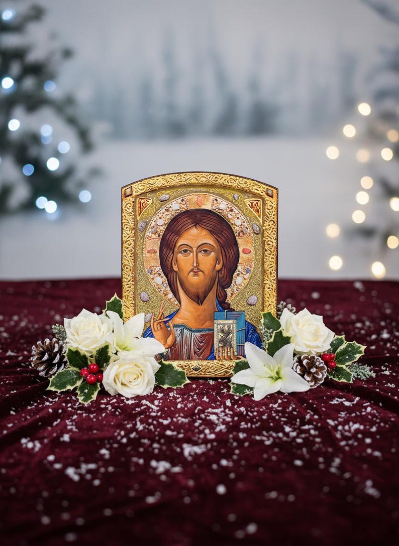 May include: An icon of Jesus Christ with a gold frame, adorned with white roses, lilies, pinecones, and holly berries. The icon is set against a burgundy velvet surface with a snowy effect, creating a festive and religious theme.