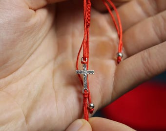 May include: Red string bracelet featuring a small, silver cross pendant adorned with clear crystals. The bracelet has a sliding knot closure and small silver beads. A simple, elegant religious accessory.