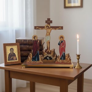 May include: A religious display featuring a wooden crucifix with a figure of Jesus, flanked by two painted figures. A framed icon and a lit candle in a brass holder sit nearby. The display is on a wooden table.