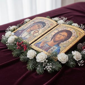 May include: Two religious icons with gold frames, depicting Jesus and Mary with child, are surrounded by a wreath of white roses, evergreen branches, and red berries. The icons rest on a deep red velvet surface.