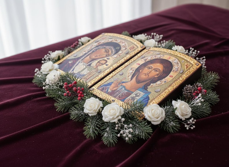 May include: Two religious icons with gold frames, depicting Jesus and Mary with child, are surrounded by a wreath of white roses, evergreen branches, and red berries. The icons rest on a deep red velvet surface.