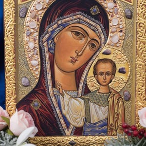 May include: A religious icon featuring a depiction of the Virgin Mary and Child. The artwork is framed in gold with ornate detailing and embellished with small stones. The Virgin Mary is depicted with a serene expression, wearing a dark red robe and blue head covering.