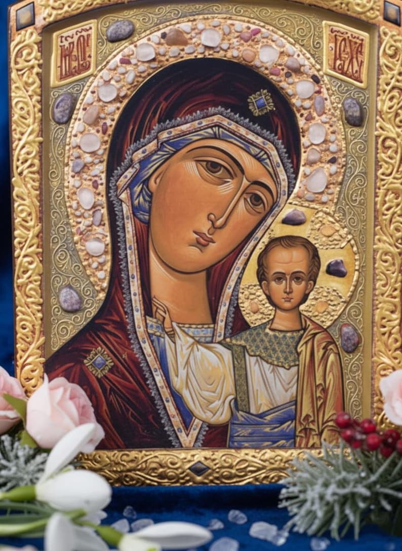 May include: A religious icon featuring a depiction of the Virgin Mary and Child. The artwork is framed in gold with ornate detailing and embellished with small stones. The Virgin Mary is depicted with a serene expression, wearing a dark red robe and blue head covering.