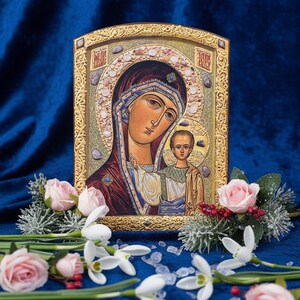 May include: An icon featuring a religious image of a woman and child, framed in gold. The icon is surrounded by pink roses, white flowers, and greenery, set against a deep blue velvet background.