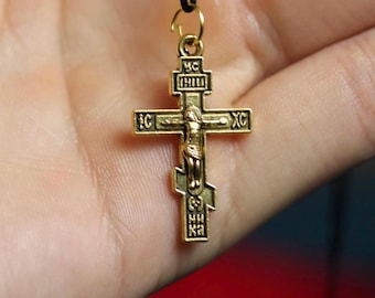 May include: Gold-colored religious cross pendant with a figure of Jesus Christ. The cross has intricate detailing and is suspended from a black cord. The cross is held in a person's hand.