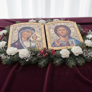 May include: Two religious icons with gold frames, depicting figures with halos, are surrounded by a wreath of evergreen branches, white roses, and red berries. The icons rest on a deep red velvet surface, creating a serene and decorative display.