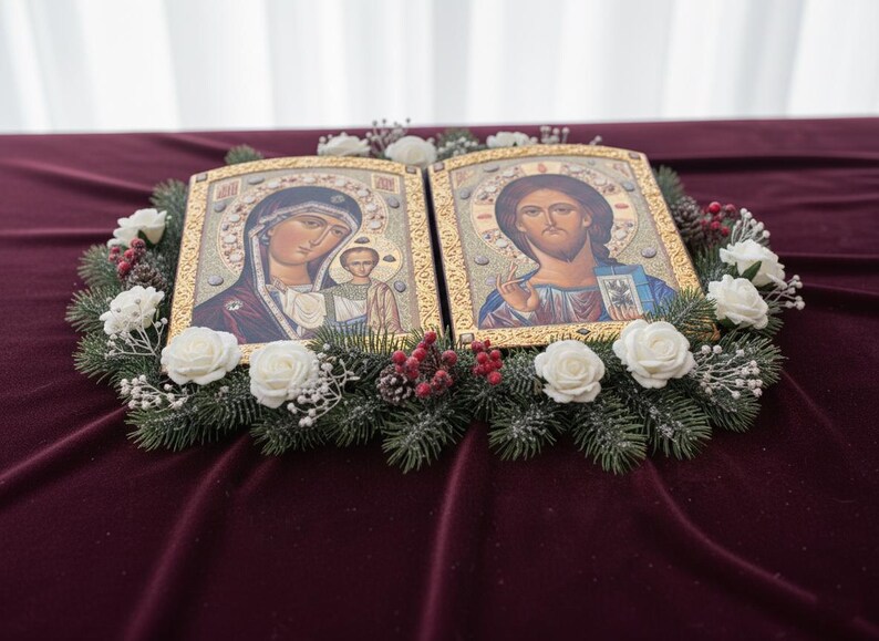 May include: Two religious icons with gold frames, depicting figures with halos, are surrounded by a wreath of evergreen branches, white roses, and red berries. The icons rest on a deep red velvet surface, creating a serene and decorative display.