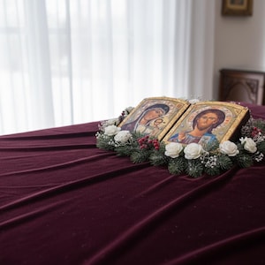 May include: Two religious icons with gold frames, depicting figures with halos, are displayed on a burgundy velvet surface. The icons are surrounded by a wreath of white roses, greenery, and red berries. The background includes a window with sheer curtains.