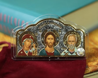 May include: A religious icon featuring three figures with gold accents and a silver background. The figures are depicted with halos and traditional religious attire. The icon is set on a red velvet surface, with a book in the background.