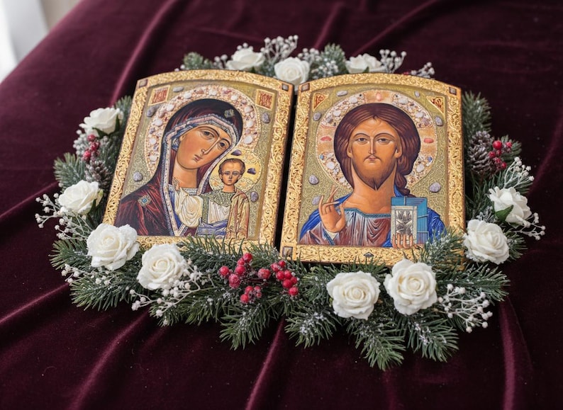 May include: Two religious icons with gold frames, depicting Mary and Jesus, surrounded by a wreath of white roses, greenery, and red berries. The icons are set against a deep burgundy background.