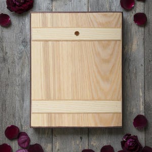May include: A light-colored wooden cutting board with a dark brown border and a small hole. The board has two light-colored horizontal stripes. Dark red rose petals and a rose are scattered around the board on a gray wooden surface.