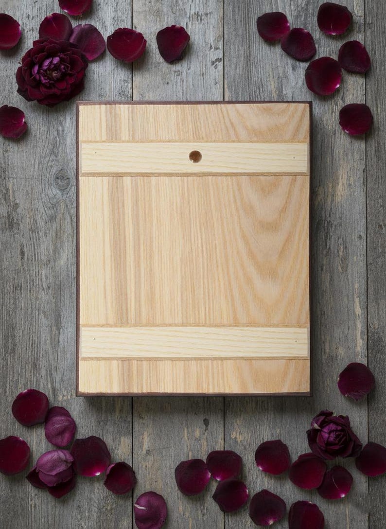 May include: A light-colored wooden cutting board with a dark brown border and a small hole. The board has two light-colored horizontal stripes. Dark red rose petals and a rose are scattered around the board on a gray wooden surface.