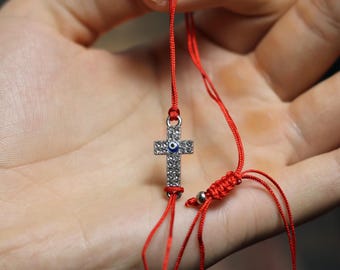 May include: A red string bracelet with a silver cross pendant. The cross is encrusted with small, clear crystals and features a blue eye in the center. The bracelet is adjustable.
