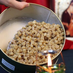 May include: A round, metal tin filled with small, light brown, cubed food items. The tin is held by a hand, with a lit candle in a green glass holder in the foreground. The tin has text on the side.