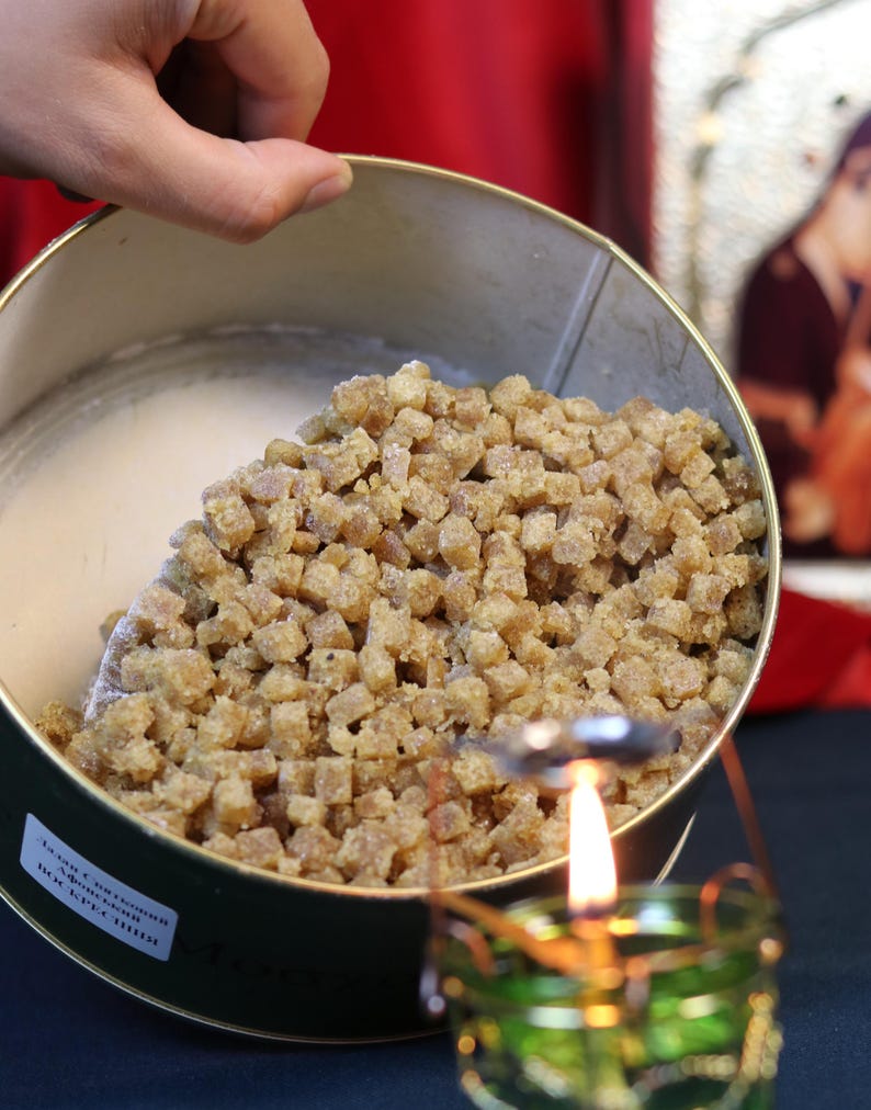 May include: A round, metal tin filled with small, light brown, cubed food items. The tin is held by a hand, with a lit candle in a green glass holder in the foreground. The tin has text on the side.