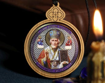May include: A wooden pendant with a colorful image of Saint Nicholas, a religious figure, surrounded by a silver and gold border. The pendant is hanging from a black cord and has a cross at the top.