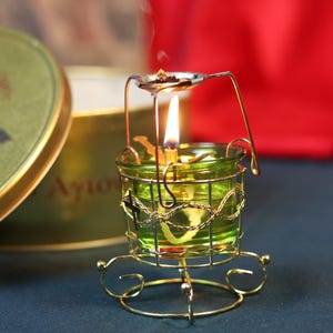 May include: A lit green glass candle holder with a gold wire cage and base. A small spoon-like holder above the candle holds incense, emitting smoke. The candle flame is bright, and the background is a blurred red and green.