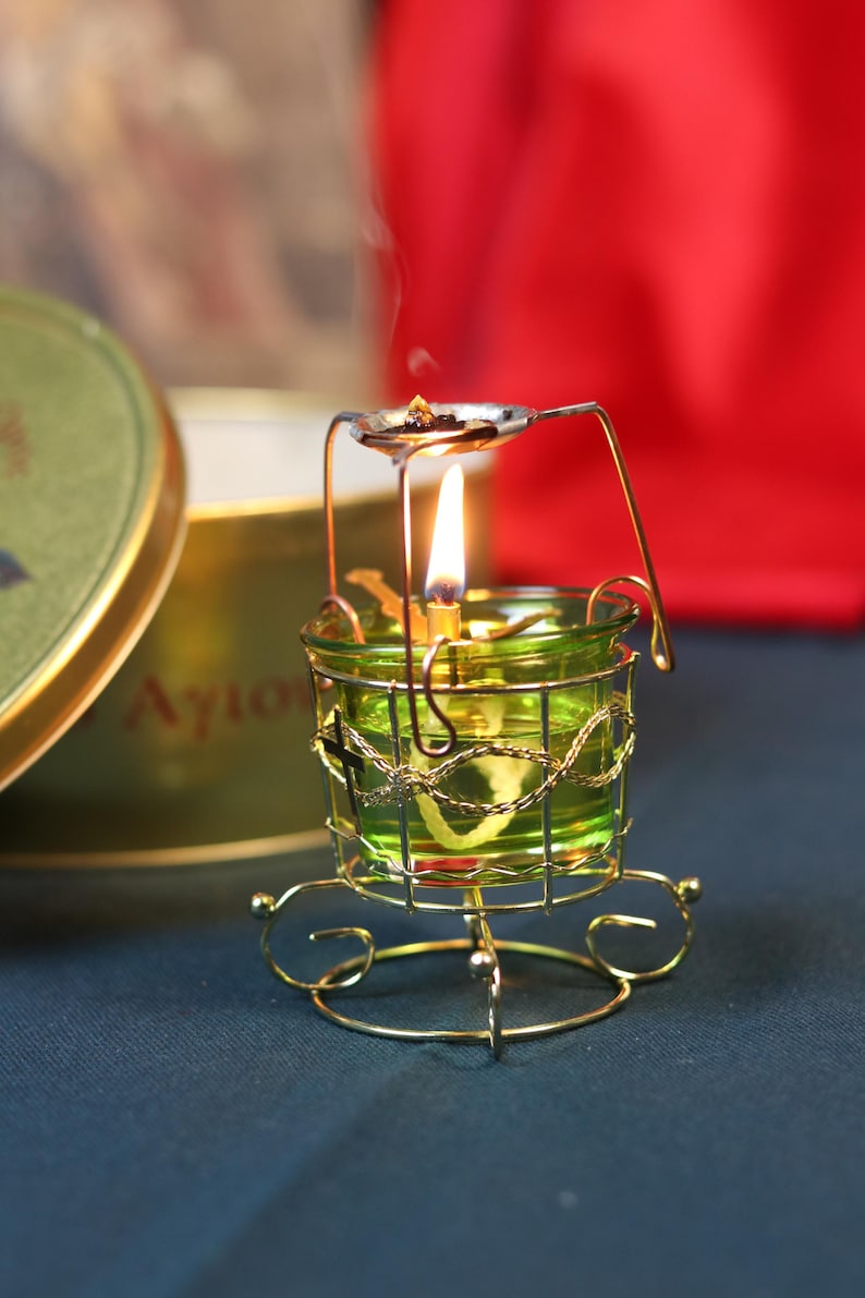 May include: A lit green glass candle holder with a gold wire cage and base. A small spoon-like holder above the candle holds incense, emitting smoke. The candle flame is bright, and the background is a blurred red and green.