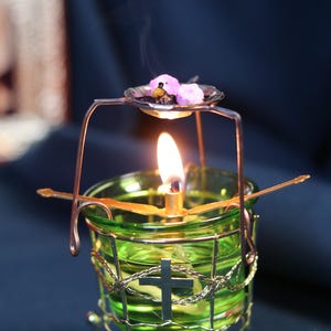 May include: A lit candle in a green glass holder with a metal cross and a decorative metal frame. The candle has a small dish above it with pink items. The flame is bright, and smoke rises from the candle.