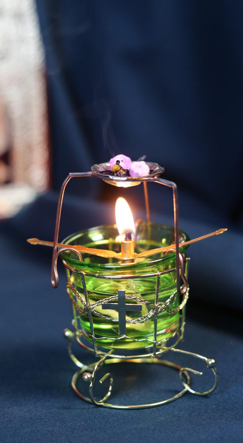 May include: A lit candle in a green glass holder with a metal cross and a decorative metal frame. The candle has a small dish above it with pink items. The flame is bright, and smoke rises from the candle.