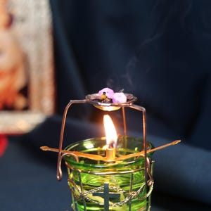 May include: A lit green glass candle holder with a cross design, surrounded by a gold wire cage. A small metal dish above the flame holds purple flowers. Smoke rises from the flame against a dark background.